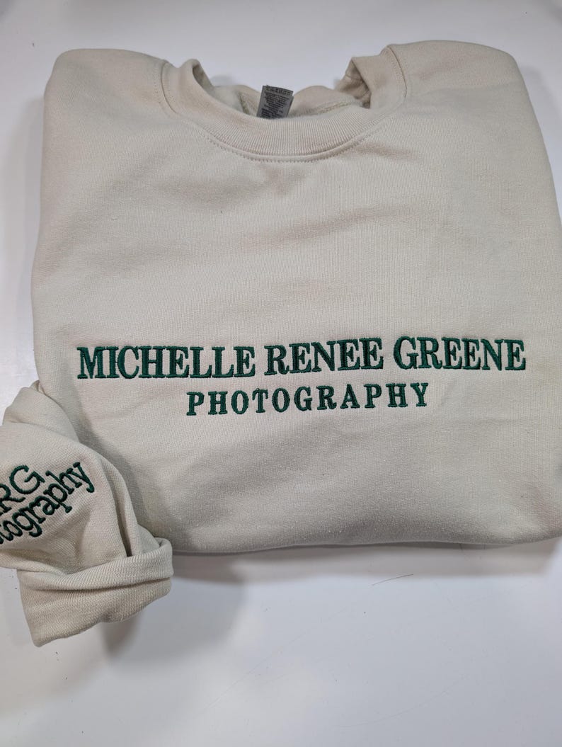 May include: Cream-colored sweatshirt with the embroidered text "MICHELLE RENEE GREENE PHOTOGRAPHY" in green. The sleeve cuff also features the embroidered letters "MRG Photography" in green.