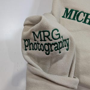 May include: Cream-colored sweatshirt with green embroidered text. The sleeve cuff displays "MRG. Photography" in a stylized font. The word "MICHAEL" is also embroidered in green on the garment.