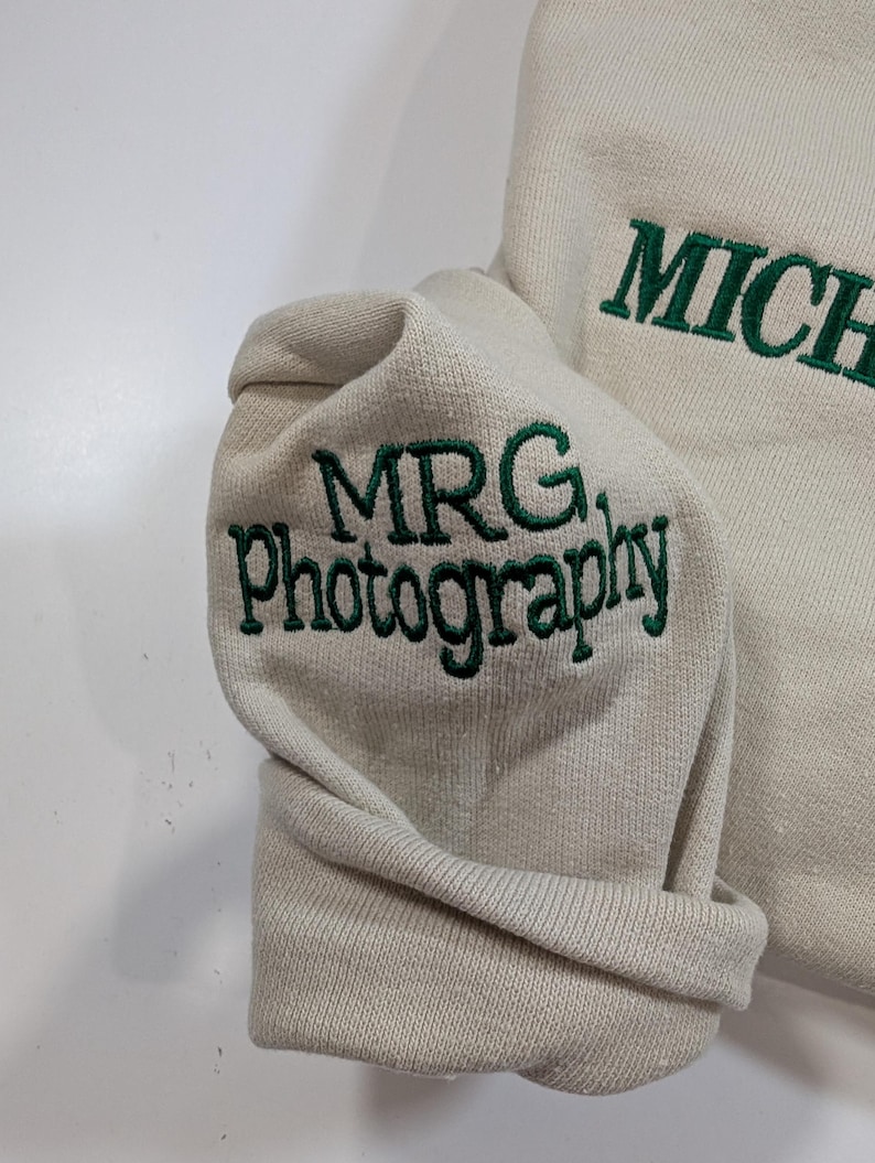 May include: Cream-colored sweatshirt with green embroidered text. The sleeve cuff displays "MRG. Photography" in a stylized font. The word "MICHAEL" is also embroidered in green on the garment.