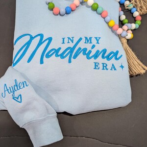 May include: Light blue sweatshirt with the text "In My Madrina Era" embroidered in blue on the front. The left sleeve has the name "Ayden" embroidered in blue with a heart.