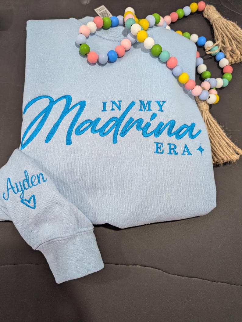 May include: Light blue sweatshirt with the text "In My Madrina Era" embroidered in blue on the front. The left sleeve has the name "Ayden" embroidered in blue with a heart.