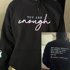 May include: Black hoodie with the text "YOU ARE enough" in pink lettering on the front and the text "dear person behind me. the world is a better place with you in it. Remember that! XX♡ the person in front of you" in pink lettering on the back.