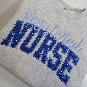 May include: A grey sweatshirt featuring the embroidered words "Registered Nurse". The word "NURSE" is in large, sequined block letters in blue, with "Registered" in a lighter blue script.