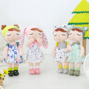 May include: Four plush dolls with different outfits and hair accessories. The dolls have closed eyes and are wearing dresses with floral patterns. One doll has bunny ears.