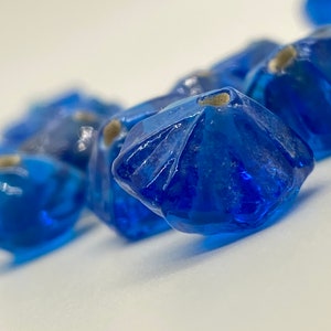 Puede incluir: Primer plano de varias cuentas de vidrio azul profundo facetadas. Cada cuenta tiene una forma única con una textura ligeramente rugosa y un pequeño agujero de color claro en la parte superior. Las cuentas están dispuestas muy juntas, creando un patrón visualmente atractivo.
