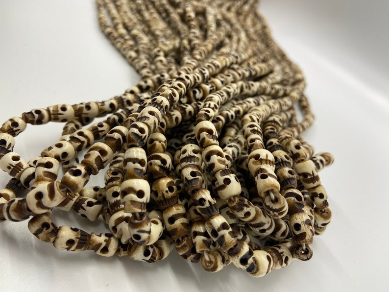 May include: A strand of white and brown skull beads. The beads are made of bone and have a carved skull design.