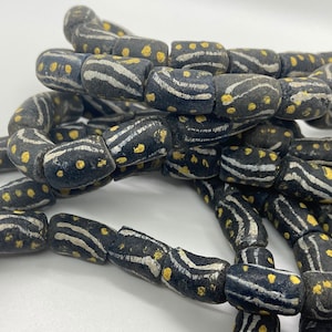 Puede incluir: Una hebra de cuentas de vidrio negro con diseños pintados en blanco y dorado. Las cuentas son cilíndricas y tienen un patrón ondulado con puntos dorados.