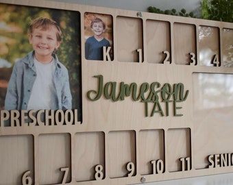 May include: Wooden photo display board with spaces for photos from preschool through senior year. Includes the name "Jameson Tate" in green script. The board has spaces for photos and the grade levels are labeled with numbers.