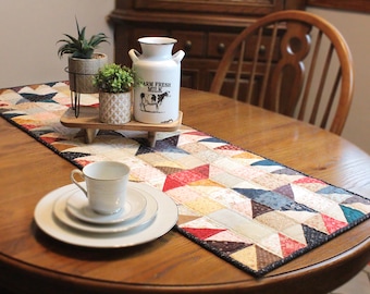 May include: A colorful patchwork table runner with a geometric pattern of triangles. The runner is on a wooden table with a white cup and saucer, a milk jug that says "Farm Fresh Milk", and two potted plants.