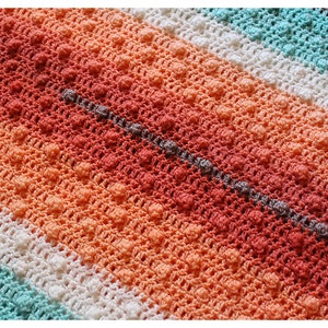 May include: A close-up of a crocheted blanket with a striped pattern in shades of coral, peach, white, and turquoise.