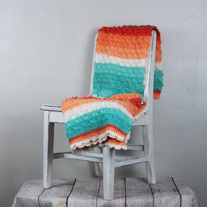 May include: A white chair with a crocheted blanket in shades of orange, white, and teal draped over it. The chair is sitting on a wooden surface.
