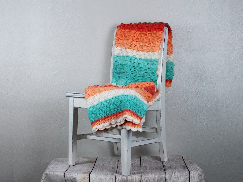 May include: A white chair with a crocheted blanket in shades of orange, white, and teal draped over it. The chair is sitting on a wooden surface.
