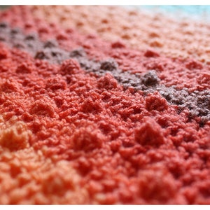 May include: Close-up of a multi-colored crocheted blanket with a textured, bumpy surface. The blanket is made with a gradient of peach, orange, red, and purple yarn.