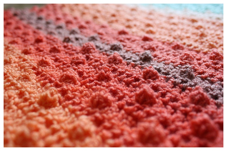 May include: Close-up of a multi-colored crocheted blanket with a textured, bumpy surface. The blanket is made with a gradient of peach, orange, red, and purple yarn.