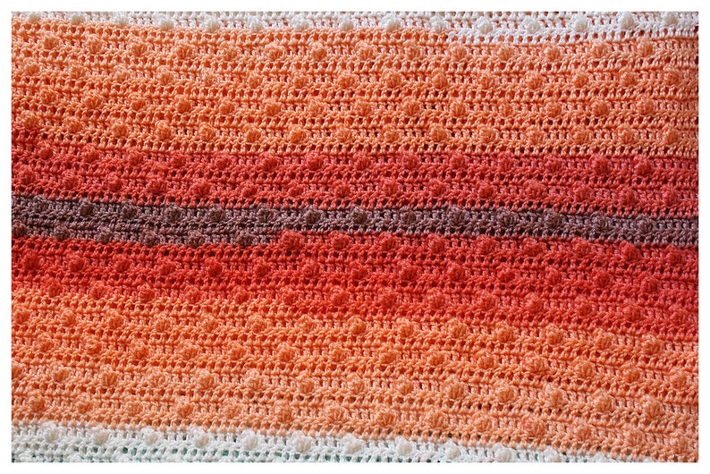 May include: A crocheted blanket with a textured pattern in shades of orange, red, and brown. The blanket has a white border.