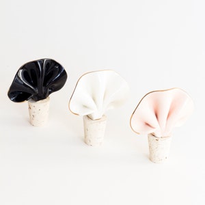 Peut inclure: Trois bouchons de bouteille en céramique de couleur noire, blanche et rose avec une bordure dorée. Chaque bouchon a la forme d'un morceau de tissu plié et repose sur une base en liège.