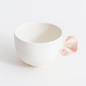 May include: A white ceramic teacup with a distinctive handle in the shape of a pink flower petal. The cup has a smooth, matte finish and a simple, modern design. Ideal for tea or coffee.