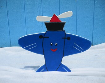May include: A whimsical, blue wooden airplane decoration with white wing accents, a red triangle, and a black hat. The airplane has a smiling face and is set against a blue wooden background and snow.