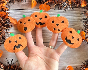 May include: Five orange felt finger puppets shaped like pumpkins with different facial expressions. The puppets are worn on a hand against a woodgrain background with orange and black garland.