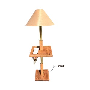 May include: A wooden table lamp with a beige lampshade. The lamp has a square base and a small shelf. The lamp's metal components are gold-colored. The lamp is plugged into an electrical outlet.