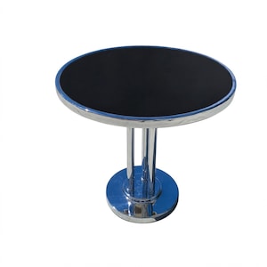 May include: A round table featuring a smooth black top surface and a polished chrome metal frame. The table is supported by a central pedestal base consisting of three vertical chrome columns that connect to a circular chrome floor plate.