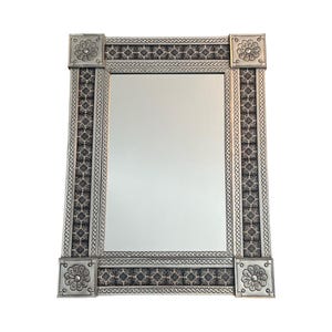 May include: Rectangular silver-colored mirror with a decorative frame. The frame features a repeating geometric pattern in black and silver, with floral corner accents. The mirror is designed for wall mounting and adds a touch of elegance to any room.