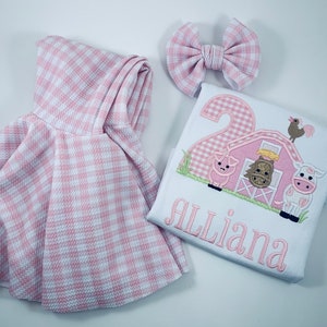May include: Pink and white gingham dress, white t-shirt with a pink gingham number 2, a pink barn with farm animals, and the name "Alliana" embroidered on the shirt.  The set includes a pink and white gingham bow.
