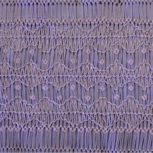 May include: Close-up of a lavender fabric with detailed smocking. The smocking forms a textured pattern of vertical lines, zigzags, and small floral accents. The fabric has a soft, matte finish.