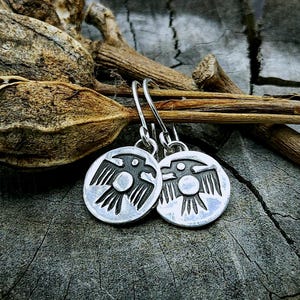 May include: A pair of silver earrings with a stylized bird design. The earrings are hanging from a wooden surface.
