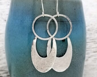 May include: Silver dangle earrings with a hammered crescent moon shape and a circle above. The earrings are displayed on a blue ceramic stand.