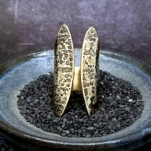 May include: A pair of gold-toned, textured, open-ended rings. The rings are placed on a bed of black pebbles in a blue ceramic bowl.
