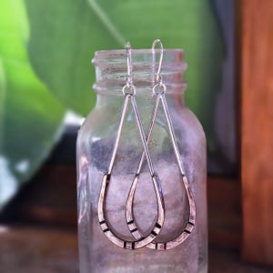 May include: A pair of silver and copper-toned teardrop earrings. The earrings have a long, slender silver top that connects to a copper-colored teardrop shape with black rectangular accents. The earrings are displayed in front of a glass jar.
