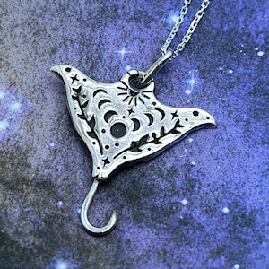 May include: A silver-colored pendant necklace featuring a stylized manta ray design. The pendant has intricate cut-out details of stars, moons, and suns. The necklace hangs on a delicate chain against a dark blue background with a starry pattern.