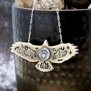 May include: A brass pendant necklace featuring a stylized eagle with celestial and mystical symbols, including a crescent moon, stars, hands, and a heart. The pendant is attached to a silver chain.