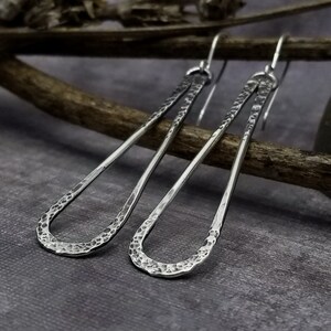 May include: A pair of silver teardrop earrings with a hammered texture. The earrings are suspended from simple hooks and have a long, slender shape. The textured surface catches the light, creating a subtle sparkle. The earrings are displayed on a wooden branch against a gray background.