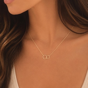 May include: A delicate gold necklace featuring two interlocking circles. The necklace is worn on a person with long, wavy brown hair and a white tank top. The jewelry is simple and elegant.
