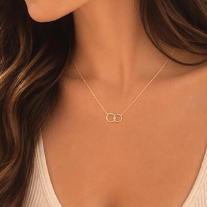 May include: A delicate gold-tone necklace featuring two small, interlocking circular rings as a central pendant. The thin chain rests against the collarbone, showcasing a minimalist design with a polished metallic finish and a simple clasp closure.