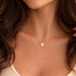 May include: A gold-toned necklace featuring a delicate chain and a small, flat, triangular pendant. The pendant is centered on the chain, which rests against the collarbone. The metal has a smooth, polished finish that reflects light.