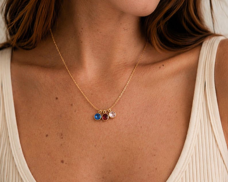 Pu&ograve; includere: Una collana dorata con tre piccoli pendenti rotondi blu, rossi e rosa. La collana &egrave; indossata su una canotta bianca a coste.