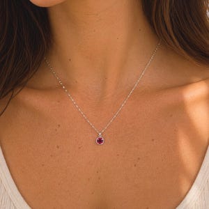 Pu&ograve; includere: Una delicata collana d'argento con un piccolo pendente rotondo color rubino. Il pendente &egrave; incastonato in una lunetta d'argento e appeso a una sottile catena. La collana &egrave; mostrata su uno sfondo neutro.