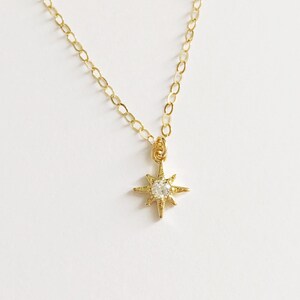 May include: A gold-toned necklace featuring a delicate chain and a small, eight-pointed star pendant. The center of the star is set with a single, clear, round-cut stone that catches the light, creating a bright, reflective focal point.