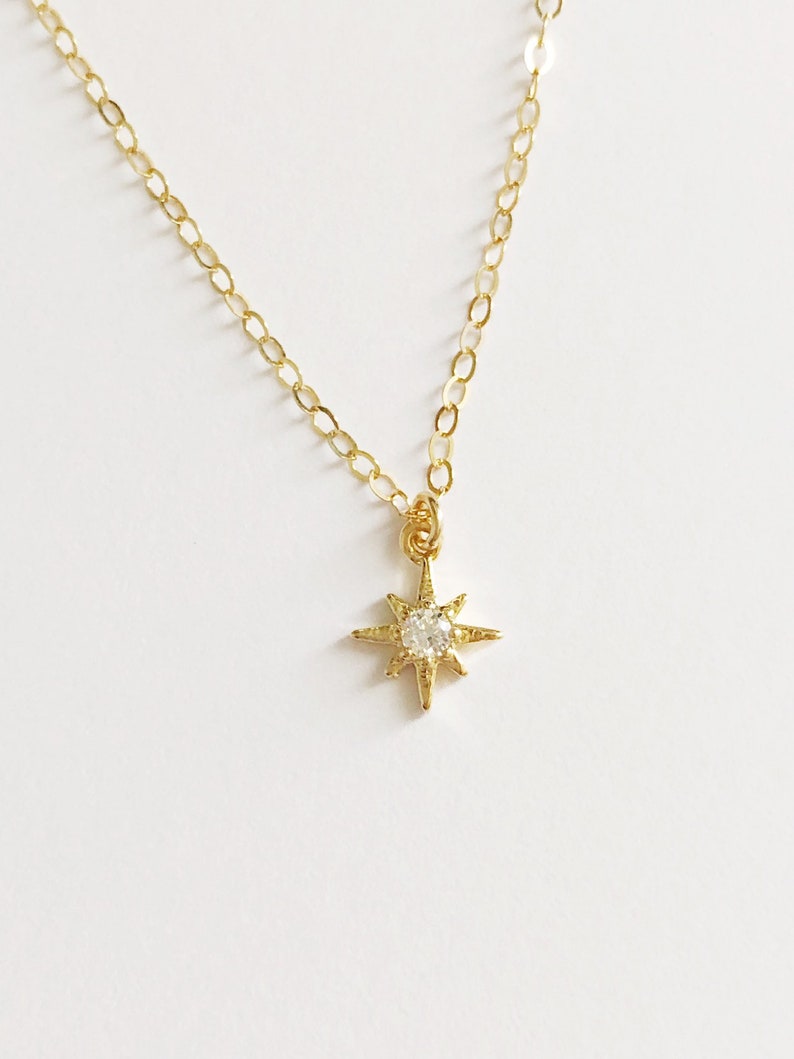 May include: A gold-toned necklace featuring a delicate chain and a small, eight-pointed star pendant. The center of the star is set with a single, clear, round-cut stone that catches the light, creating a bright, reflective focal point.