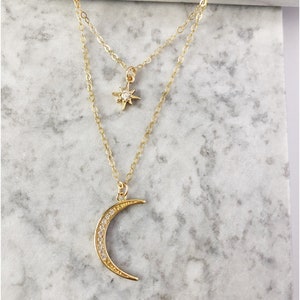 May include: A gold-toned layered necklace featuring two delicate chains. The shorter chain holds a small, four-pointed star charm with a central clear stone, while the longer chain features a crescent moon pendant encrusted with small clear stones.