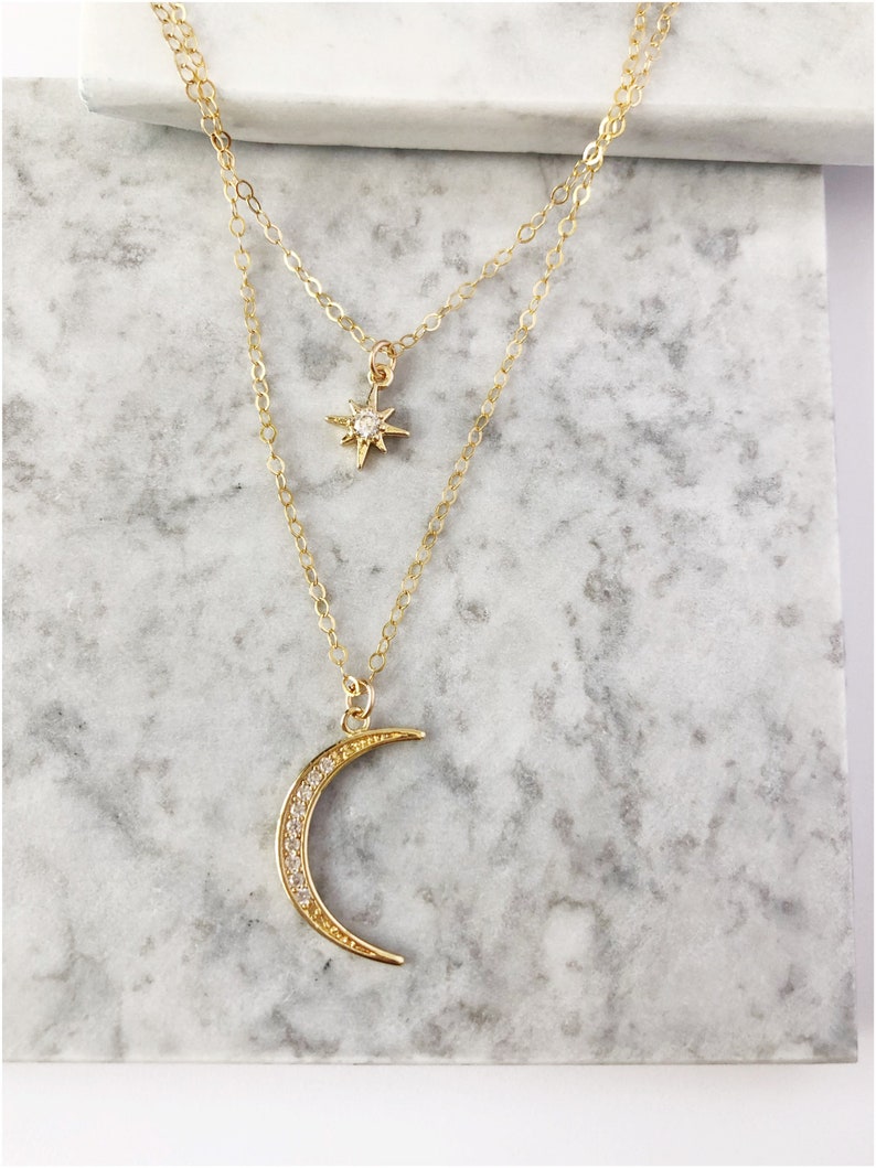 May include: A gold-toned layered necklace featuring two delicate chains. The shorter chain holds a small, four-pointed star charm with a central clear stone, while the longer chain features a crescent moon pendant encrusted with small clear stones.