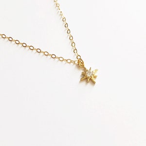 May include: A gold-toned necklace featuring a delicate chain and a small, star-shaped pendant. The pendant has a textured, multi-pointed design with a single, clear, round-cut stone set in the center, reflecting light against the metallic finish.