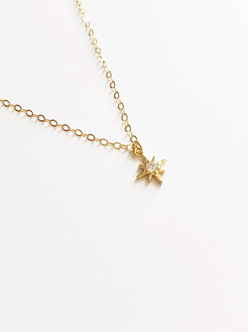 May include: A gold-toned necklace featuring a delicate chain and a small, star-shaped pendant. The pendant has a textured, multi-pointed design with a single, clear, round-cut stone set in the center, reflecting light against the metallic finish.