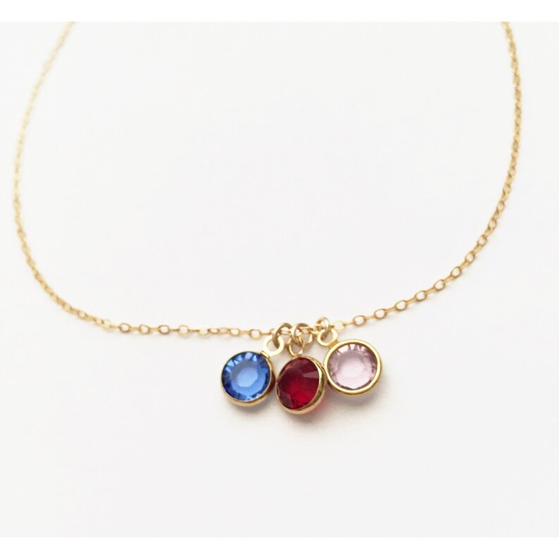 Pu&ograve; includere: Una delicata collana color oro con tre pietre preziose rotonde blu, rosse e rosa. Le pietre preziose sono sospese da una sottile catena, creando un design semplice ma elegante. La collana &egrave; posta su uno sfondo bianco.