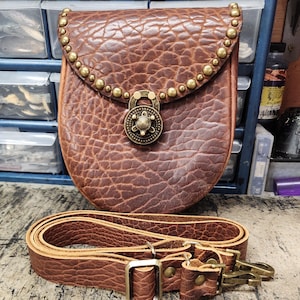 May include: A brown leather bag with a textured surface, adorned with brass studs and a decorative lock. Includes a matching adjustable strap with metal hardware. The bag is designed for carrying essentials.