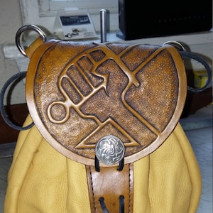 Könnte beinhalten: Ein gelber Lederbeutel mit einer braunen Ledertasche, die ein erhabenes Prägemuster einer Faust mit einem Schwert zeigt. Der Beutel hat einen schwarzen Kordelzug und einen silbernen Metallknopf.