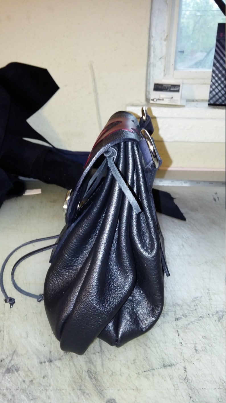 May include: A black leather bag with a drawstring closure and a red and black strap. The bag is folded and has a silver buckle.
